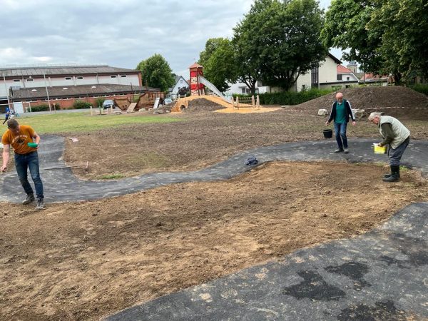 11.07.2022: Heute wurde die Fläche für die Blühwiese in der Mitte der Asphaltbahn vorbereitet: Geharkt, Steine aufgesammelt und eingesät. Bald blühen hier Wildblumen für Hummeln, Bienen, Schmetterlinge & Co. Im Hintergrund wächst bereits ganz leicht der neue Rasen. In der nächsten Woche werden Tore, Ballfangzaun und Tischtennisplatte wieder aufgestellt.